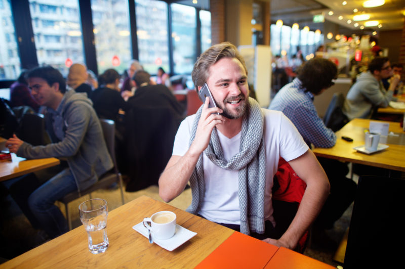 Lifestyle-Fotografie: In einem gut besetzten Café an einer Universität sitzt ein junger Mann an einem der Tische und telefoniert mit seinem Smartphone.