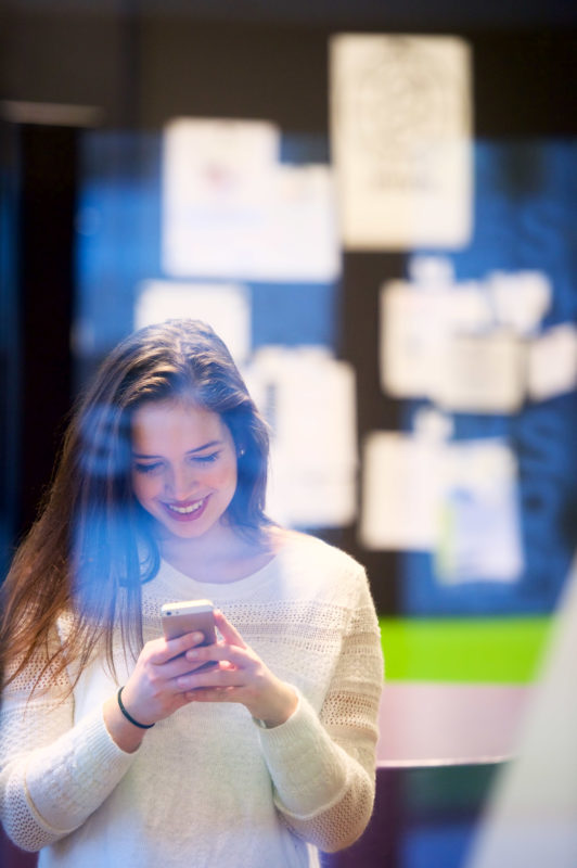 Industriefotografie: Lifestyle-Fotografie: Eine Studentin schaut im Universitätsgebäude lachend auf ihr Smartphone.