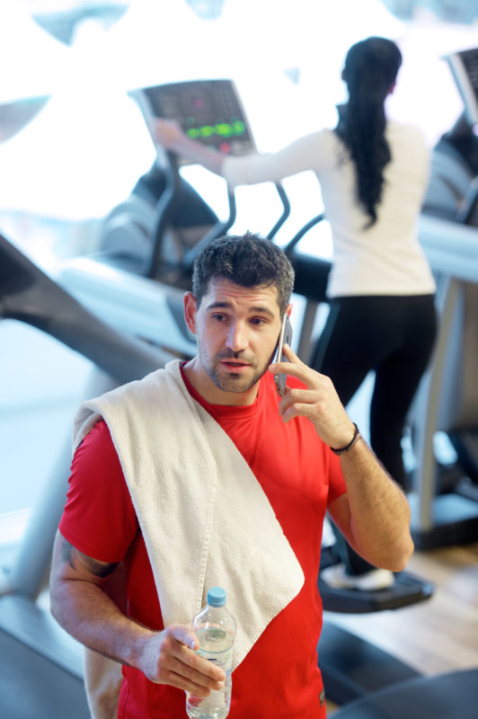Lifestyle-Fotografie: Ein Mann telefoniert in einem Fitness-Studio mit seinem Mobiltelefon. Im Hintergrund trainiert eine junge Frau auf einem Laufband.