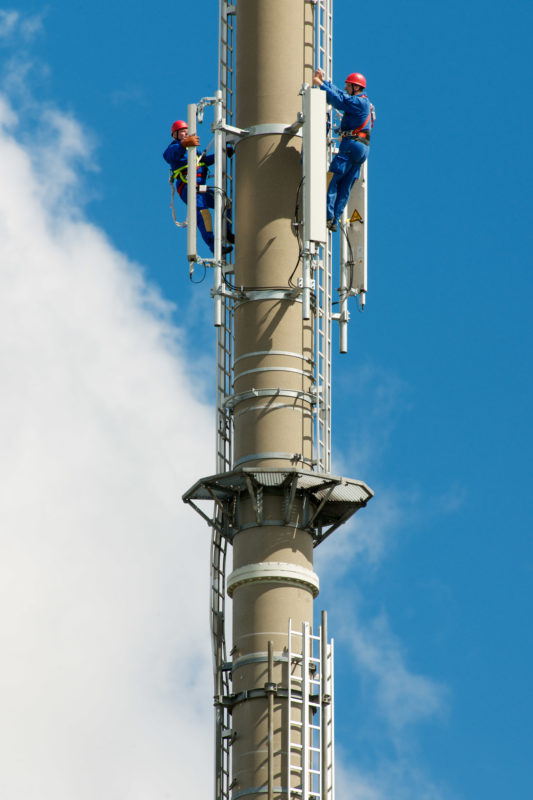 Industriefotografie: Zwei Monteure sind auf einen hohen Sendemast geklettert, um dort angebrachte LTE-Mobilfunk-Antennen zu warten. Im blauen Himmel sieht man vorbeiziehende weiße Wolken.