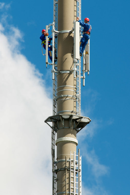 Industrial photography: Two fitters climbed a high transmission mast to maintain LTE mobile phone antennas installed there. In the blue sky you can see passing white clouds.