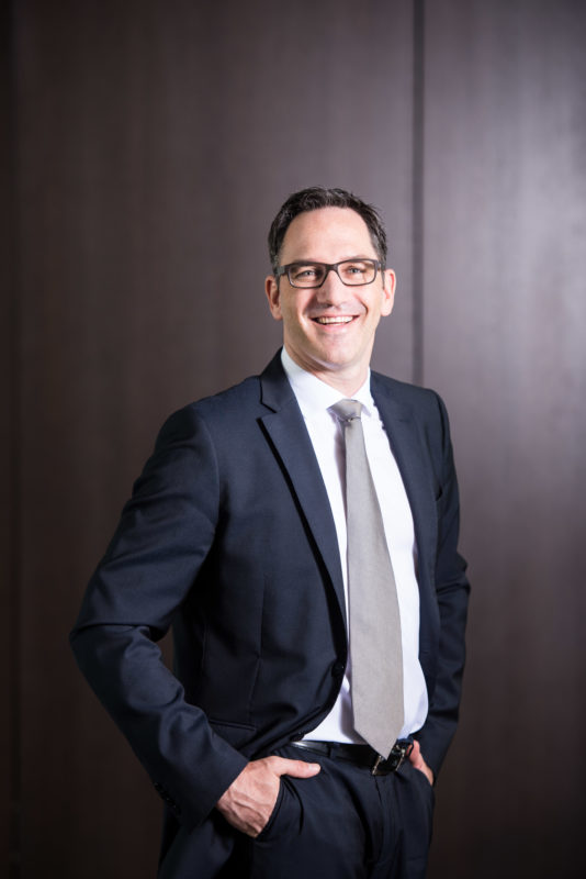 Managerportrait: A male manager stands loosely with his hands in his pockets in a suit and a tie in front of a wall clad in dark wood. The image is illuminated from the right with a large reflector so that light and shadow are accentuated.