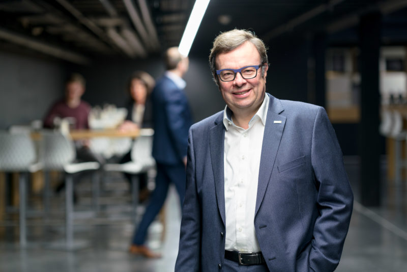 Managerportrait: Ein Manager steht in einem dunklen Flur an dem an der Decke eine Lichtleiste verläuft, die ihn anleuchtet. Im Hintergrund sieht man weitere Mitarbeiter und Mitarbeiterinnen. Das Bild ist bei wenig Licht mit offener Blende fotografiert.