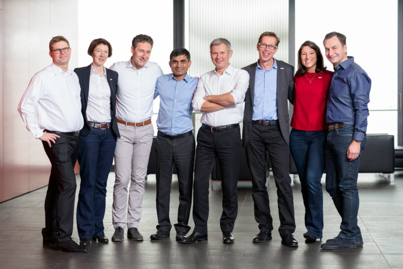 Gruppenfoto: Gemeinsames Ganzkörperportrait von Führungskräften auf dem Gang in einer Firma. Sie tragen bewusst keine Krawatten oder sind sogar im Hemd. Die Atmosphäre ist locker und entspannt.