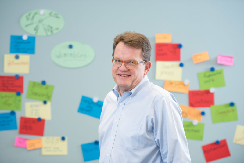 Managerportrait: A manager in a conference room in front of a notice board with many colourfully inscribed notes. He is in his shirt and is not wearing a suit or tie and smiles loosely into the camera. The picture is illuminated with a flash unit. Everything is bright and friendly.