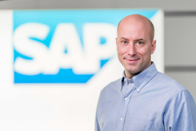 Managerportrait: A manager stands in front of his company logo. The background and him are separately illuminated with several flashes so that everything looks bright and friendly. He