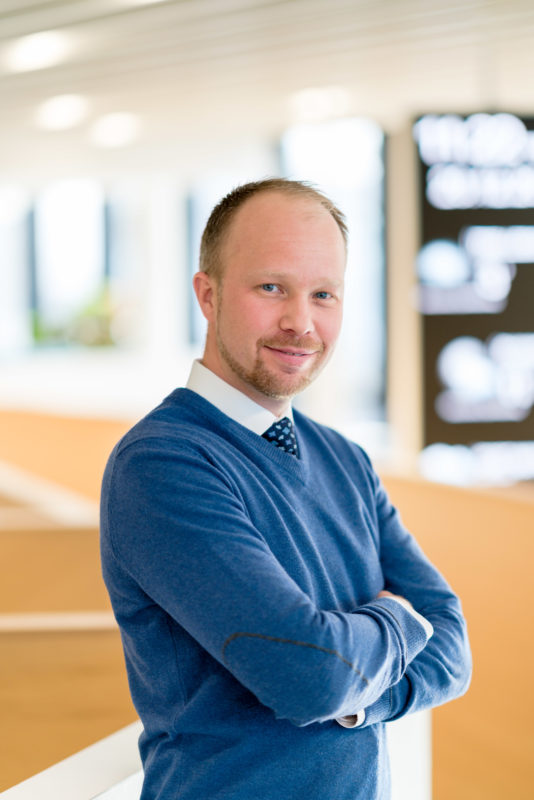 Managerportrait: Mit den Armen verschränkt steht ein Manager im Foyer seiner Firma. Im Hintergrund unscharf sieht man das helle Holz des Treppenaufgangs und ein leuchtendes senkrecht montiertes Display. Er trägt einen blauen Pullover, ein weisses Hemd und eine Krawatte.