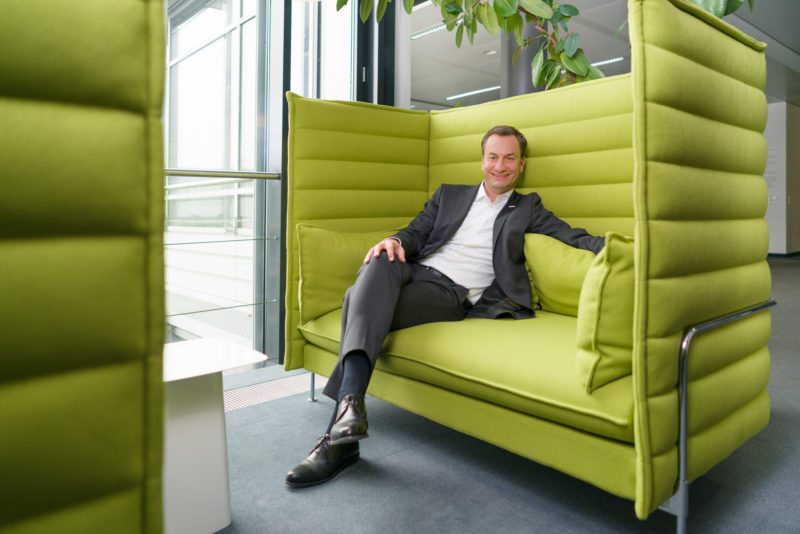 Managerportrait: Ein Manager sitzt bequem in einer Art Sofa, dessen hohe Rückenlehne ihn gegen die Umgebung abschirmt. Im Hintergrund sieht man Pflanzen und ein Fenster. Er trägt Anzug ohne Krawatte. Das natürliche Licht ist zusätzlich mit Blitzlicht aufgehellt.