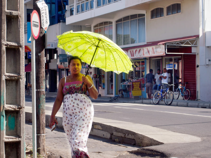 travel photography:  Mauritius: A woman with a green parasol walks on the street of a small town. Behind her is a clothing fashion store.