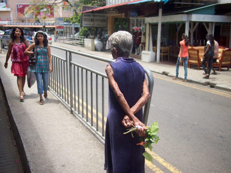 Reisefotografie: Mauritius: Eine ältere Frau mit Kräutern in der Hand zwischen jungen Damen auf einer Hauptstraße.