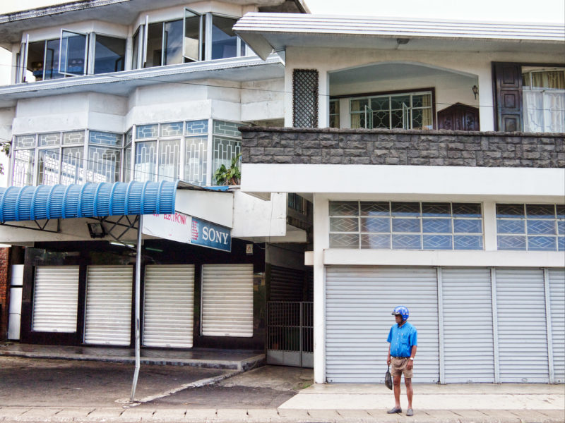 Reisefotografie: Mauritius: Ein Mann wartet am Straßenrand mit Helm auf seinem Kopf darauf, dass er auf dem Motorrad mitgenommen wird.