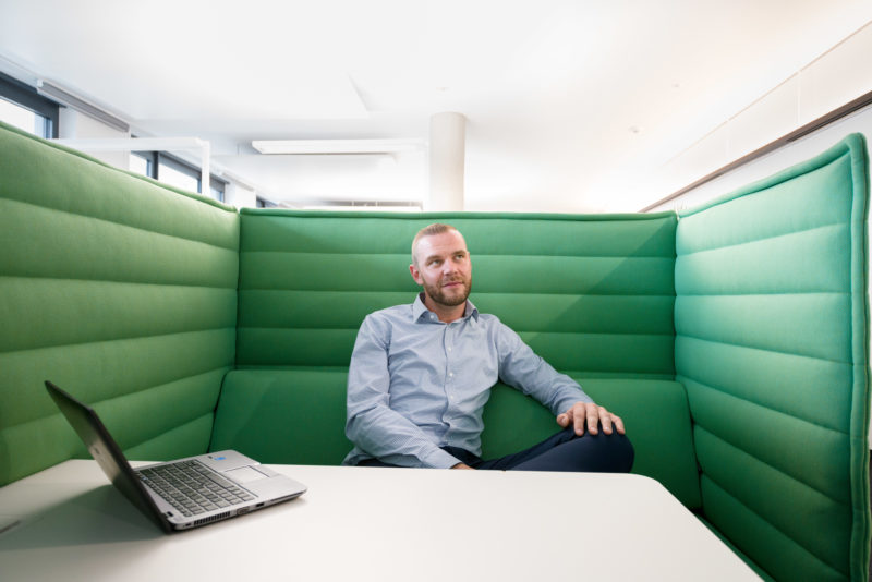 Mitarbeiterfotografie: Ein Mitarbeiter sitzt in einem modernen Büro in einer mit grünem Stoff bezogenen Besprechungsecke.