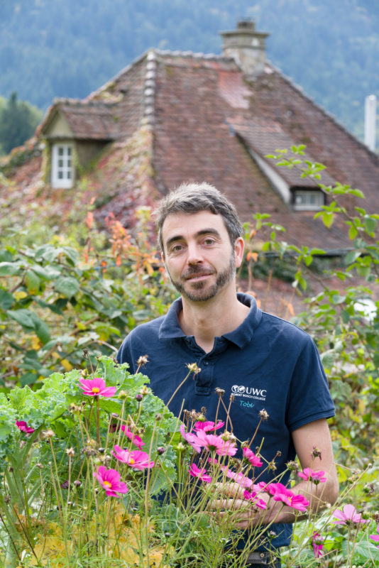 Mitarbeiterfotografie: Ein Mitarbeiter eines Colleges ist im hauseigenen Lehrgarten umgeben von Grün. Vor ihm wachsen Pflanzen mit rosa Blüten.