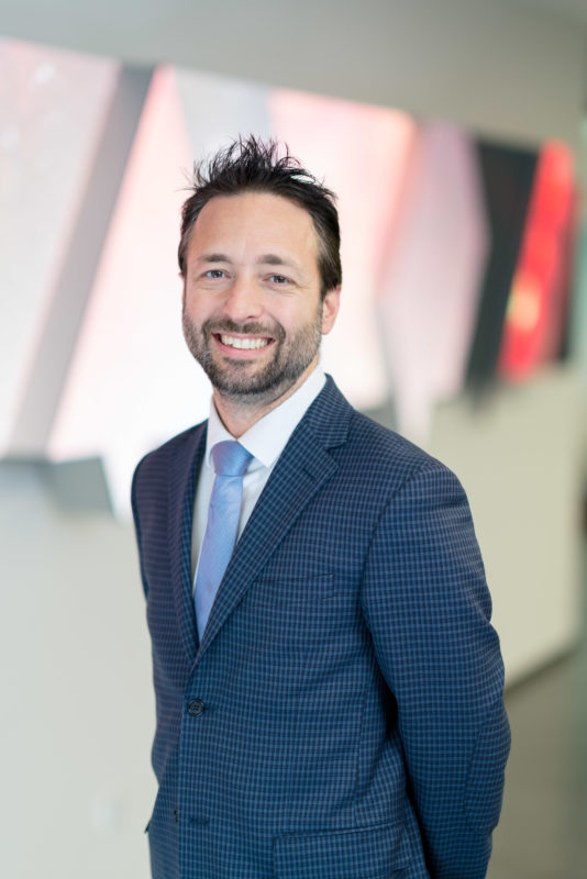 Managerportrait: A young employee of a company in front of a wall of displays in the hallway of his company. He wears a blue, slightly plaid suit and a light blue tie. He smiles into the camera. The photo is taken with a lightweight super bright telephoto lens with the aperture open.