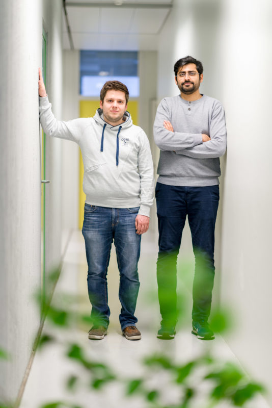 Wissenschaftsfotografie an einem Physikalischen Institut der Universität Stuttgart: Zwei Mitarbeiter auf dem Flur . Im Vordergrund sind grüne Pflanzen in dem ansonsten kahlen, weissen Raum.