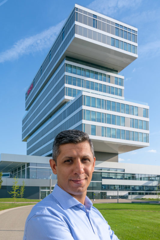 Employees photography: An employee proudly stands in front of the research building of his company. The building rises very modern and impressive into the blue sky.