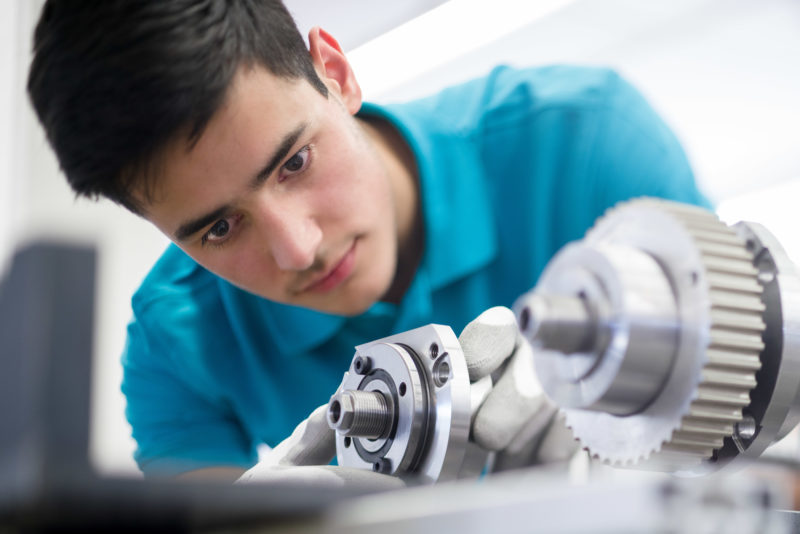 Employees photography: A young employee of a mechanical engineering company concentrates on the shiny metal gear axles in front of him. He wears work gloves and the light blue work shirt of his company.