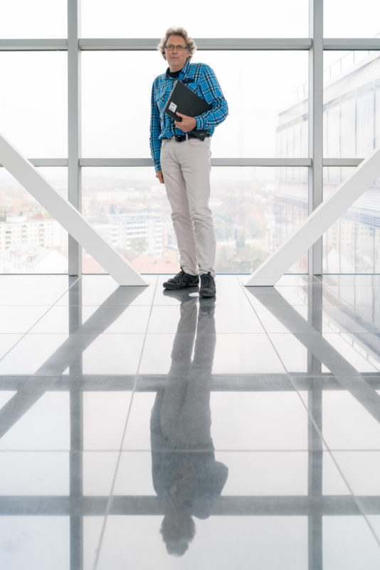 Mitarbeiterfotografie: Ein Mitarbeiter einer Softwarefirma auf einem Flur im Hochhaus des Firmengebäudes. Die großen Glasflächen geben den Blick auf die Stadt frei während der Mann sich in den glänzenden Bodenfliesen spiegelt.