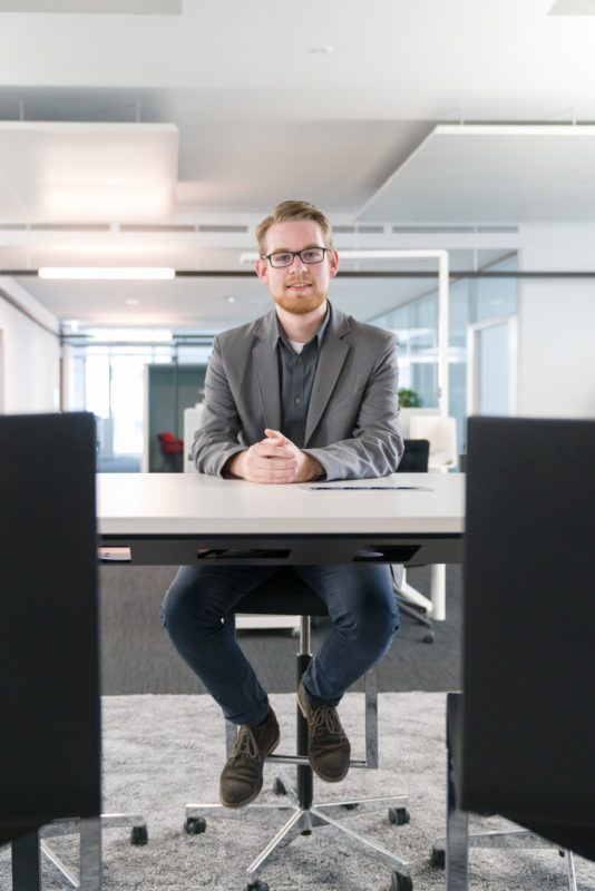 Mitarbeiterfotografie: Ein Mitarbeiter sitzt in einem modernen Büro auf einem Hocker an einem hohen Tisch und lächelt in die Kamera.