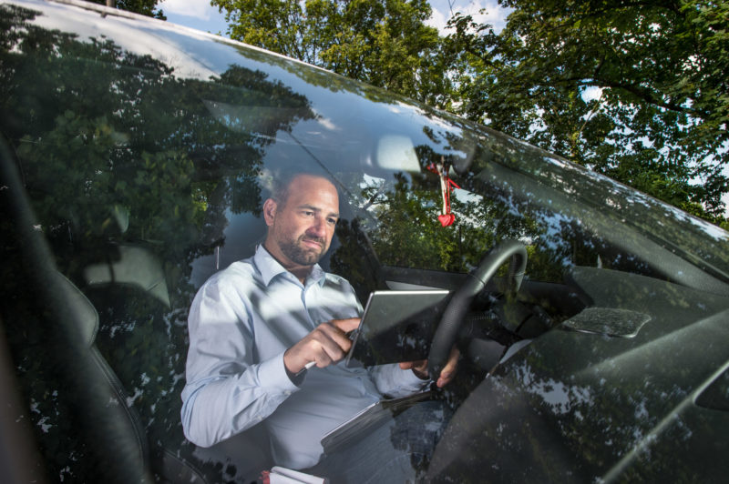 Employees photography: An employee of a telecommunications company who travels a lot works in his car sitting on a computer tablet. The trees along the parking lot are reflected in the windshield.