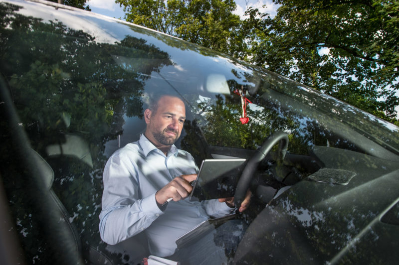 Mitarbeiterfotografie: Ein Mitarbeiter einer Telekommunikationsfirma, der viel unterwegs ist, arbeitet in seinem Auto sitzend auf einem Computertablet. In der Frontscheibe spiegeln sich die Bäume entlang des Parkplatzes.