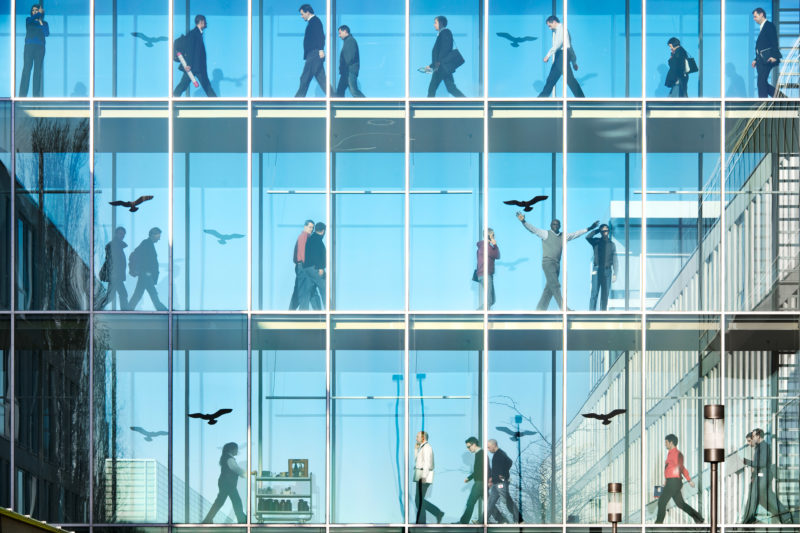 Architekturfotografie: Blick auf eine Firma und den gläsernen Verbindungsgängen in denen mittags auf allen Stockwerken Mitarbeiter unterwegs sind.