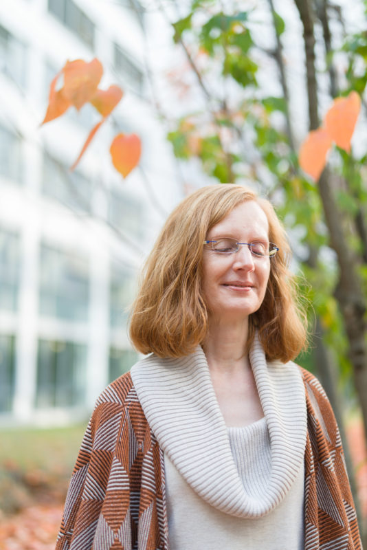 Employees photography: An employee stands with her eyes closed in the garden between two company buildings. The plants, their red hair and their clothes harmonize in colour and create a calm unity.