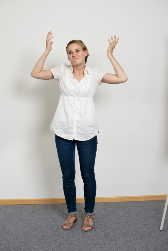 Mitarbeiterfotografie: Eine Mitarbeiterin steht auf dem typischen Teppichboden vor der weißen Wand eines Büros und macht eine Geste des Nichtwissens.