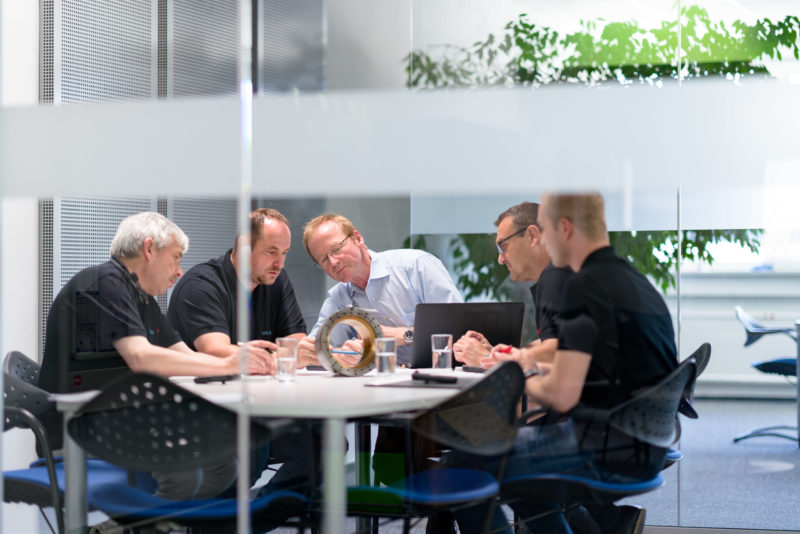 Industriefotografie: Mitarbeiter sitzen im verglasten modernen Besprechungsraum an einem Tisch, um sich über Komponenten einer neuen Maschine abzustimmen.