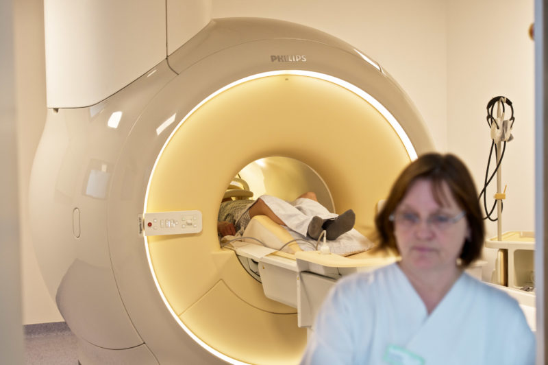 Medizinfotografie: Blick in den MRT-Raum eines Kreiskrankenhauses. Ein Patient wird während der Untersuchung auf einer Liege durch die röhrenförmige Öffnung des Magnetresonanztomograph hindurchgefahren. Im Vordergrund sieht man eine unscharf abgebildete Mitarbeiterin.