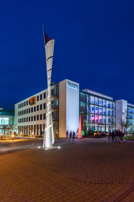 Architectural photography: Nightly illuminated building of the Fraunhofer Institute for Industrial Mathematics ITWM in Kaiserslautern with a high helically formed metallic plastic in front of it.