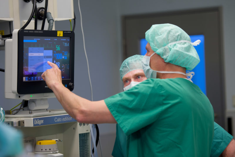 Healthcare photography:  In the operating theatre, a doctor operates the touch screen of an anaesthesia device while a surgeon talks to him.