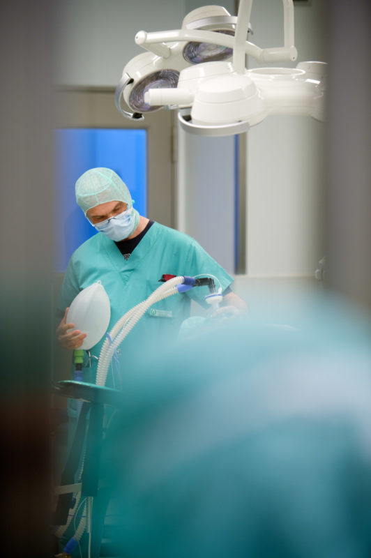 Medizinfotografie: Ein Narkosearzt leitet im Operationssaal die Narkose bei einem Patienten ein. Er hat einen Beatmungsbeutel abgenommen um die Schläuche des Beatmungsgeräts an den Tubus anzuschließen.