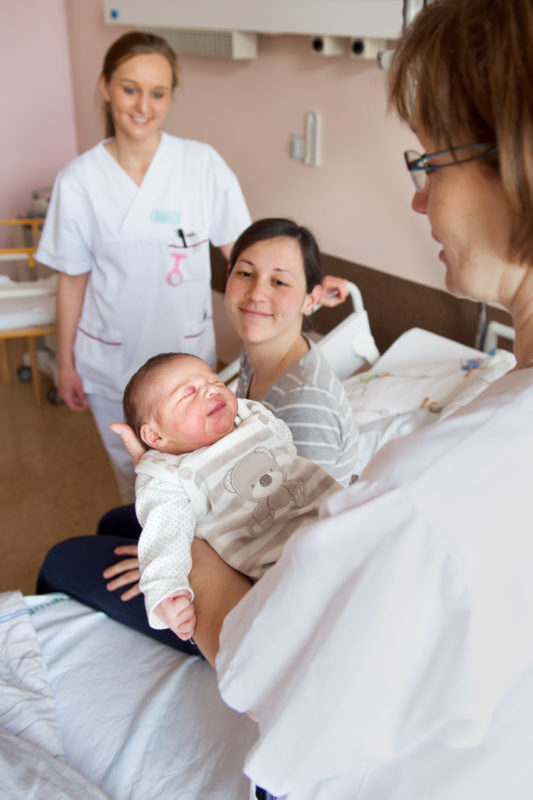 Medizinfotografie: Eine junge Mutter schaut stolz auf ihr Neugeborenes das ihr von einer Stationsschwester gebracht wird.