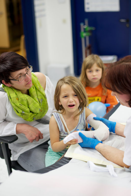 Medizinfotografie: Ein kleines Mädchen bekommt in der Notaufnahme eines Kreiskrankenhauses einen Gips um ihren gebrochenen Arm. Ihre Mutter versucht sie zu trösten.