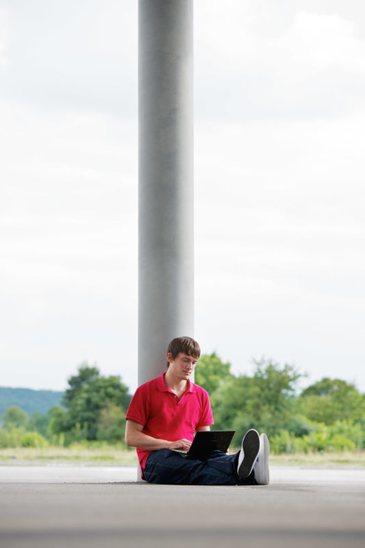 Industrial photography: A young man sits outside on the floor with his notebook and works.