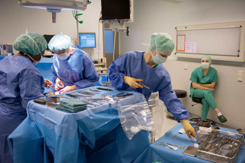Medizinfotografie: Während die Operateure am geöffneten Patienten arbeiten, kümmert sich eine Assistentin um das Besteck. Im Hintergrund sitzt eine angehende Ärztin, um den Verlauf der OP zu beobachten.