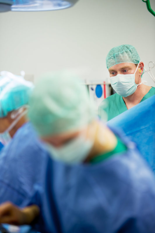 Healthcare photography: A doctor observes the course of an operation. Like everyone else in the operating room, he wears a mask and a green hood.