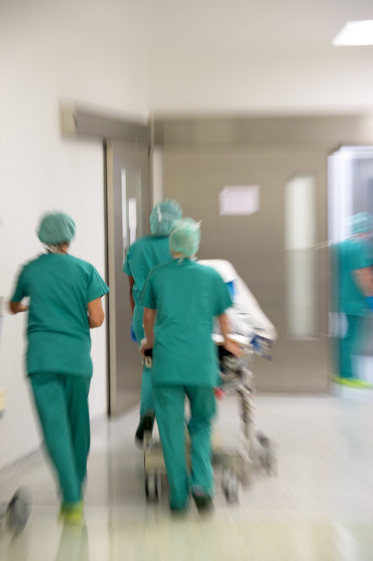 Medizinfotografie: Nach der Operation wird ein Patient im Bett von drei Mitarbeitern aus dem Operationssaal geschoben.