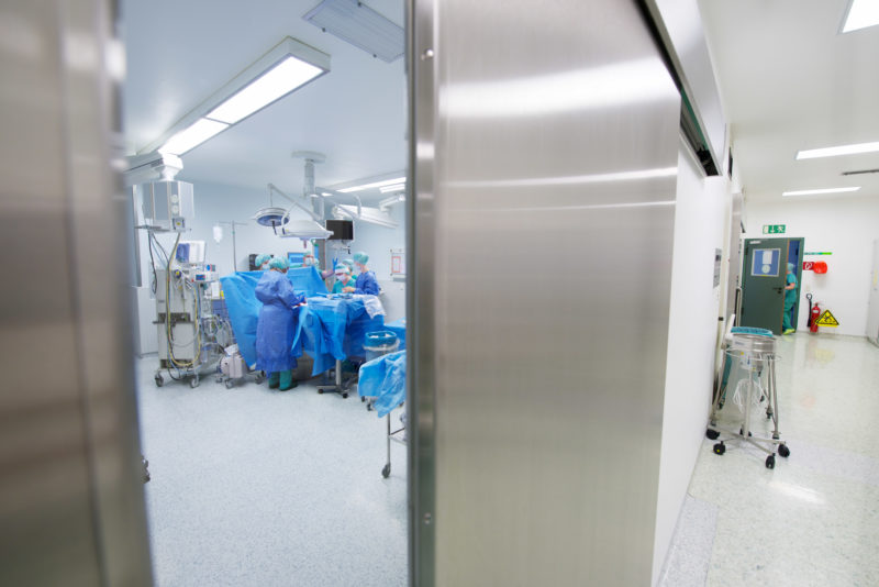 Medizinfotografie: Foto vom Flur durch die automatische Edelstahltür hinein in den Saal  in dem gerade eine Operation läuft.