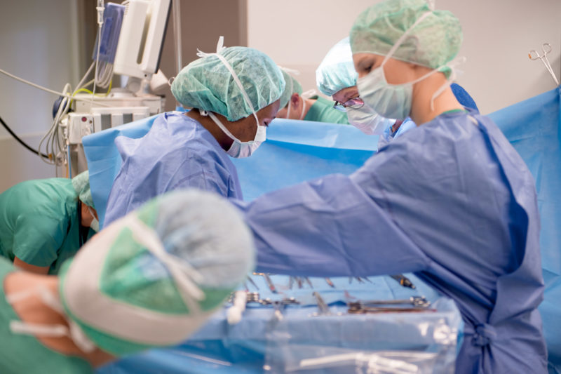 Healthcare photography: In the operating theatre of a district hospital, doctors and staff are standing around the operating table to perform an operation. On the photo you can see the heads with hoods and mouthguards.