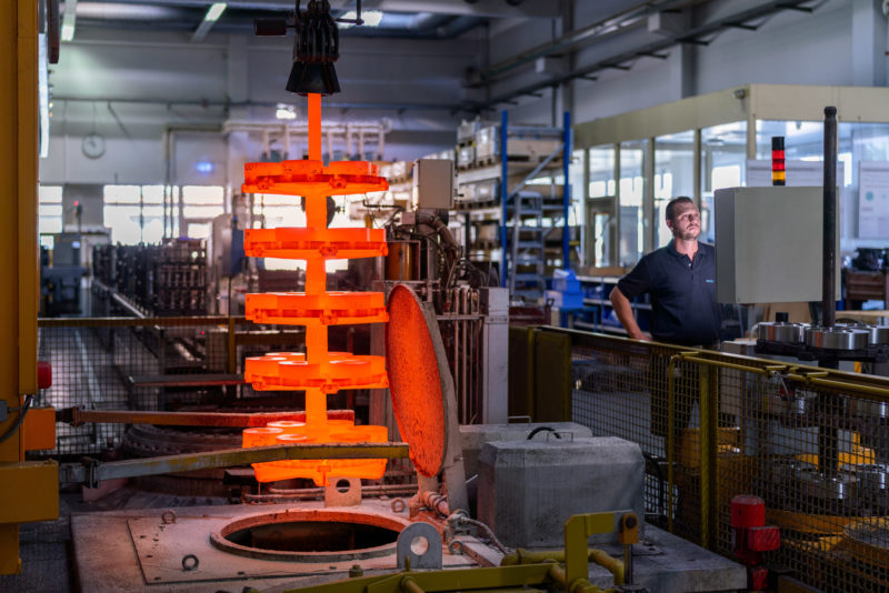 Industriefotografie: Per Kran hebt ein Mitarbeiter zum Glühen gebrachte  in oranger Farbe leuchtende rohe Metallteile aus dem Ofen.