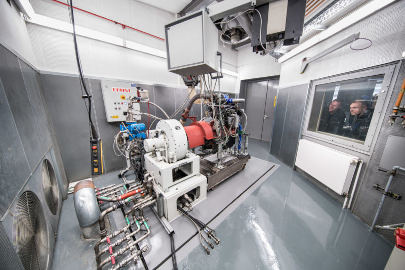 Industrial photography: Panorama photo of the interior of an engine test bench. The motor is connected to a braking device so that its power can be measured. Two employees look at the combustion engine from the steering position through the double safety glass pane.