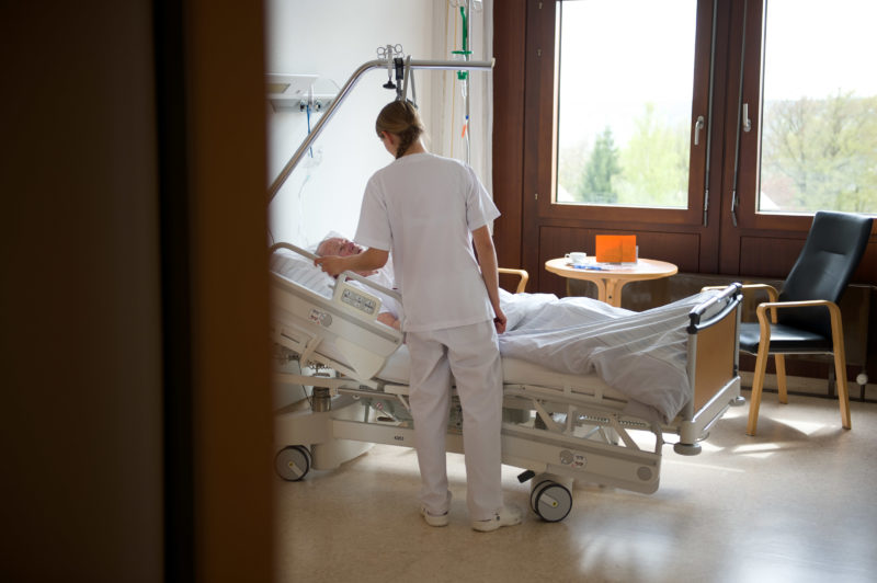 Medizinfotografie: Blick in ein Krankenzimmer eines Krankenhauses. Ein älterer männlicher Patient liegt in dem modernen verstellbaren Bett und spricht mit einer Pflegerin.