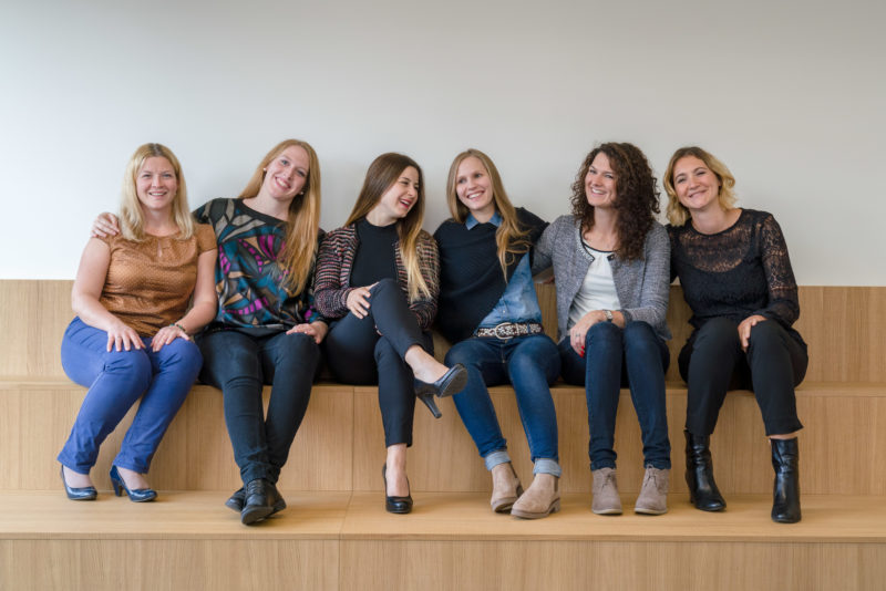 Group photo: Six members of a human resources department sit together on a bench in their company and are happy.