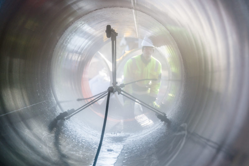 Industrial photography: Internal cleaning of a steel pipe for a pipeline. You can see the rotating water nozzle as it drives through the pipe. Behind it is an employee with a white helmet and a bright yellow safety vest.