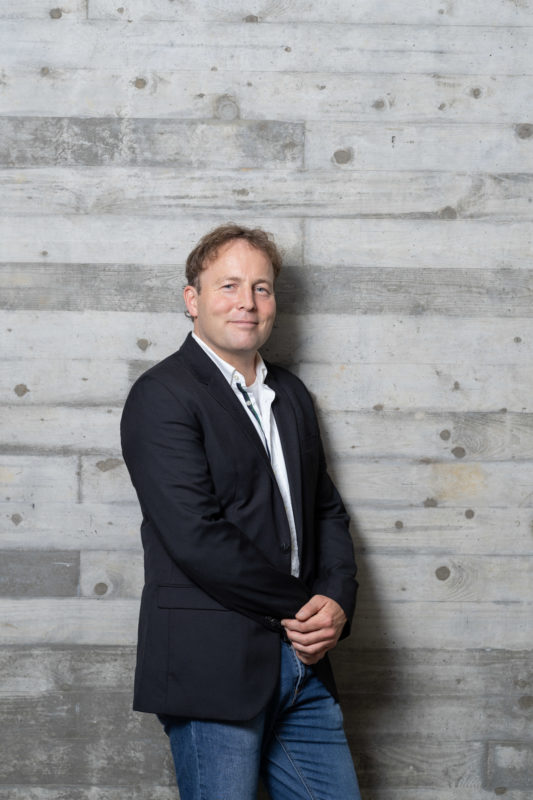 Executive portrait: A man in a casual jacket casually leans in front of a strongly structured grey concrete wall. He smiles into the camera.