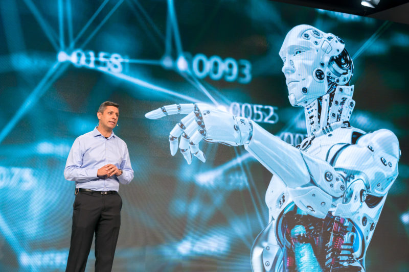 PR-Fotografie: Pressefotos auf der Pressekonferenz der Robert Bosch GmbH: Ein Mitarbeiter bei einem Statement. Im Hintergrund wird ein humanoider Roboter gezeigt.