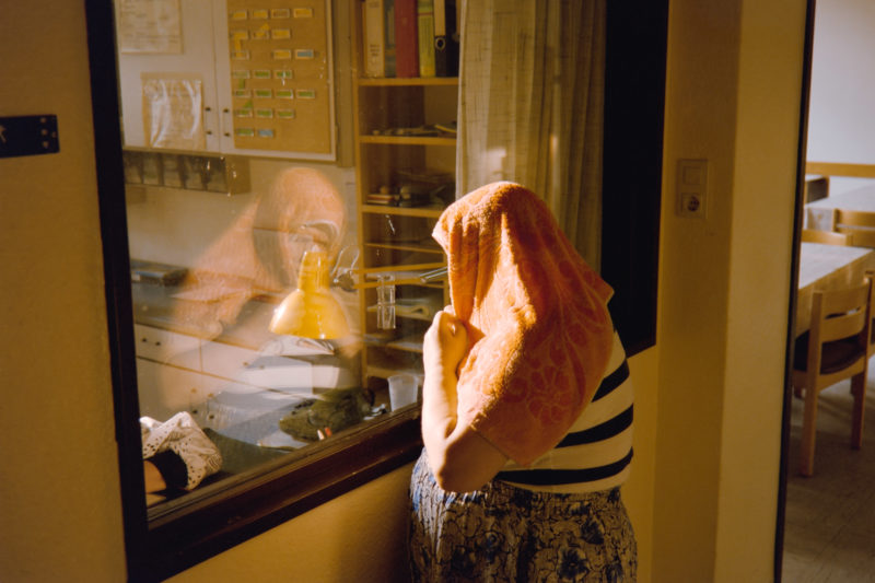 editorial photography at a psychiatric ward: A female patient wears a towel as head protection and looks through a pane of bullet-proof glass into the staff