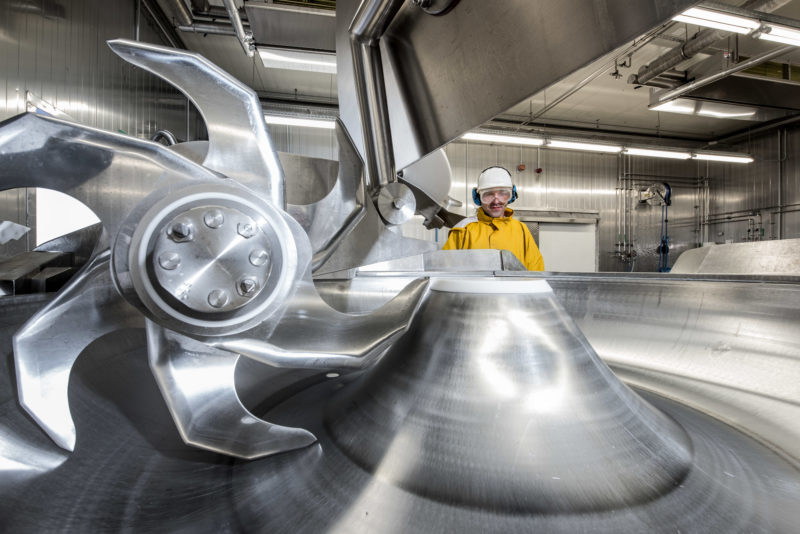 Industriefotografie: Ein Mitarbeiter eines Reinigungsunternehmens für Lebensmittelhersteller in gelbem Hygieneanzug mit Schutzbrille und Helm schaut auf einen großen Fleischkutter, der jede Nacht in der Nutzungspause extrem gründlich gereinigt wird.